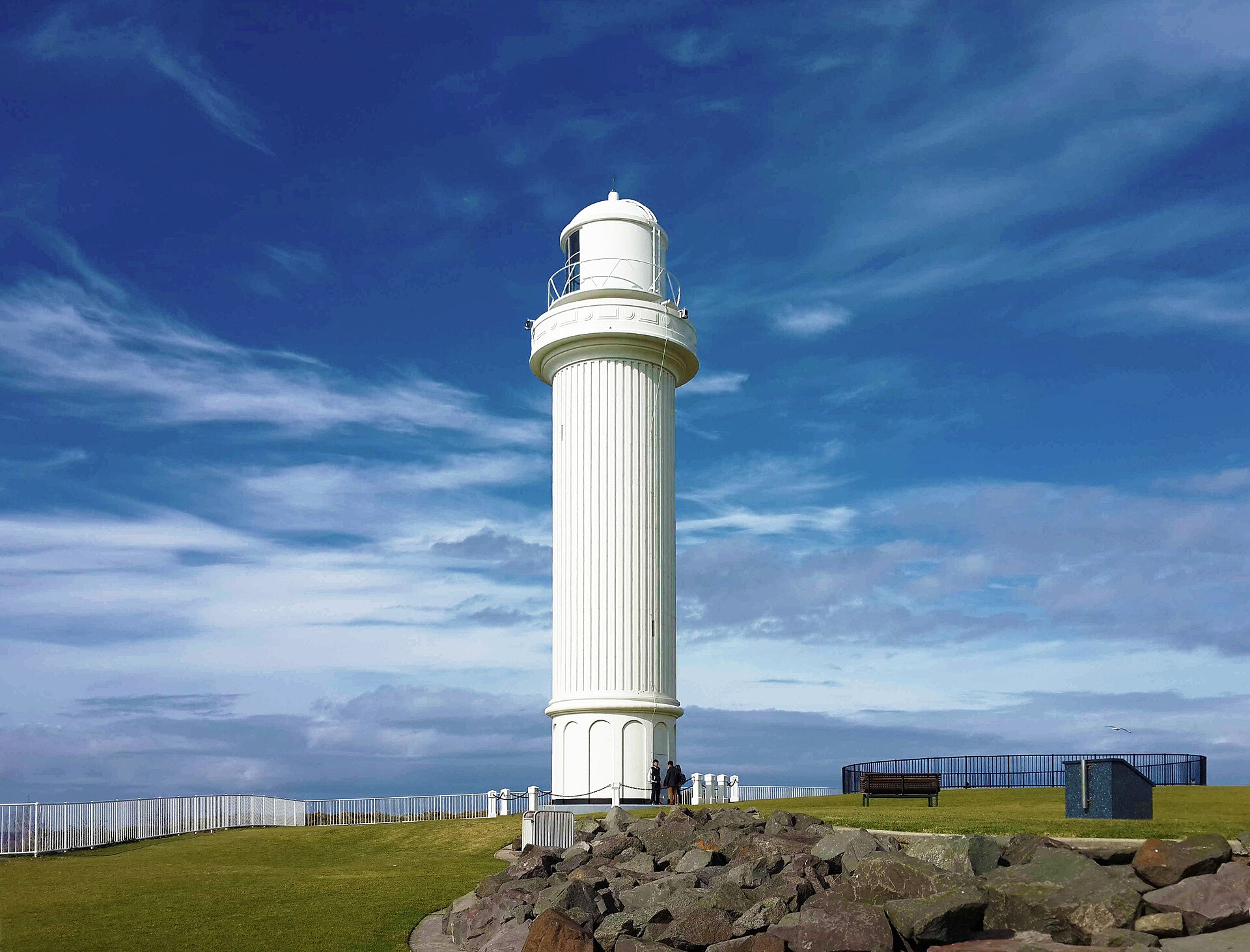 The tall white fluted tower of Wollongong Head Lighthouse on the grassy headland at Flagstaff Point, with rock armour in front and a deep blue sky